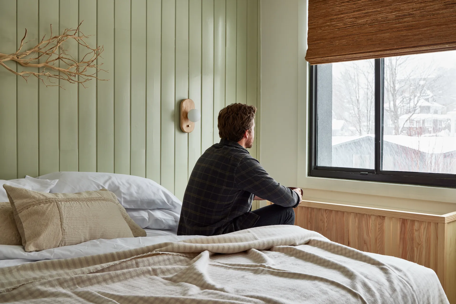 A man sits on the edge of a lush bed in a mint colored room and looks out the window at the fresh snow.