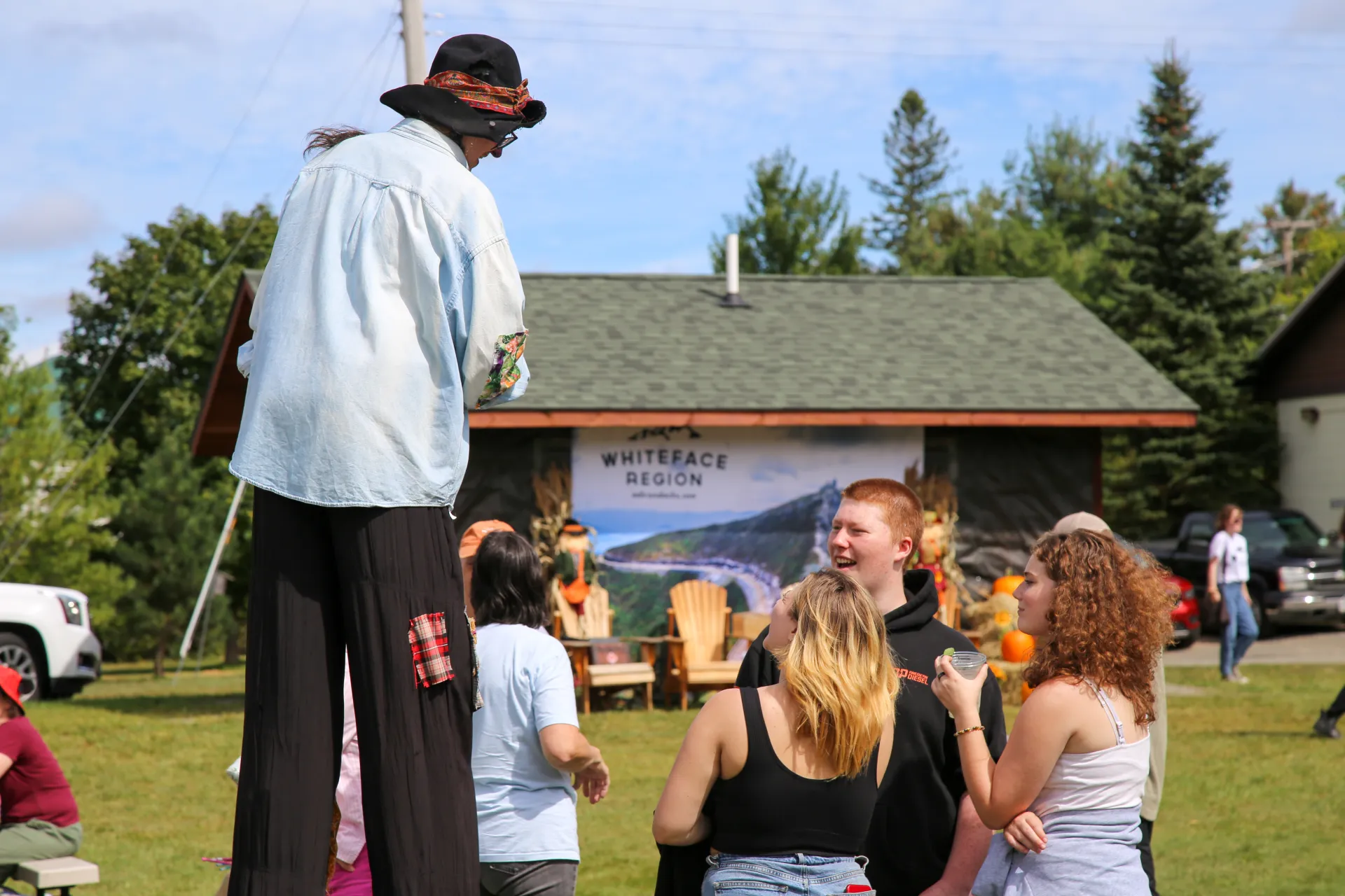 Collection of youths stand in a group talking to a woman dressed as a scarecrow on stilts
