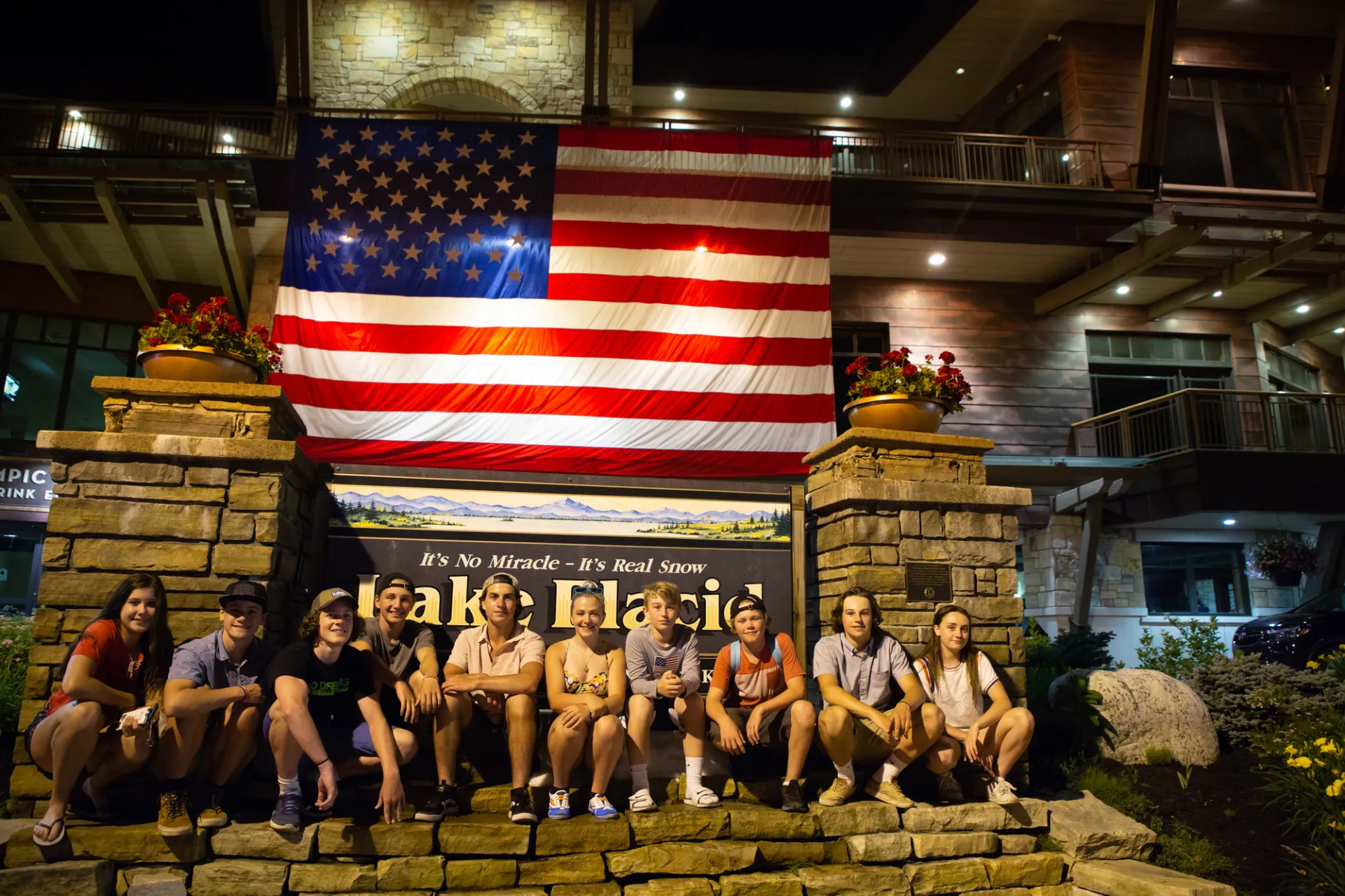 A group of young people sit in front of the Lake Placid welcome sign which has been decorated with a large American flag.