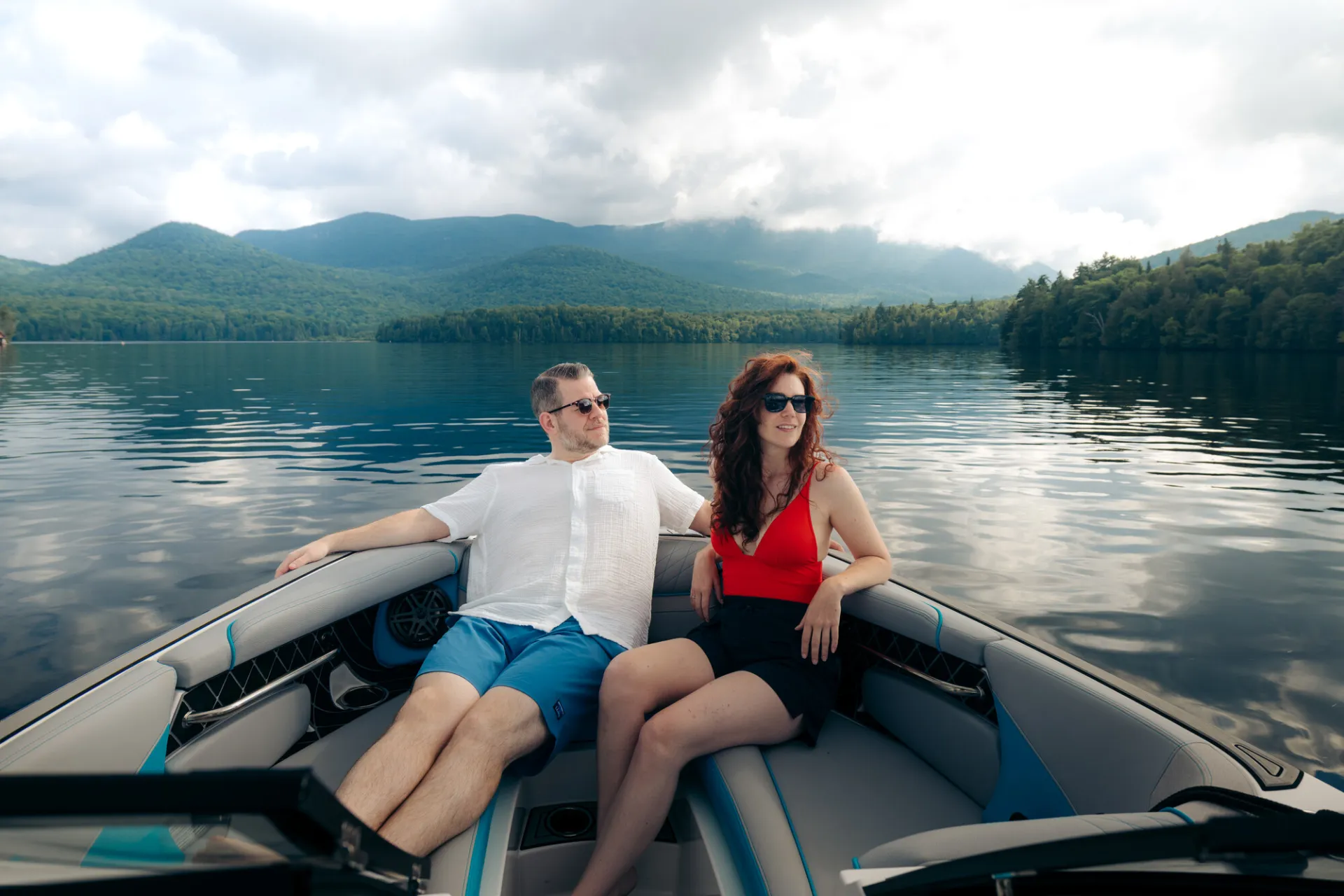 Man in white shirt and blue shorts sits next to woman in red bathing suit and shorts on back of motor boat on Lake Placid