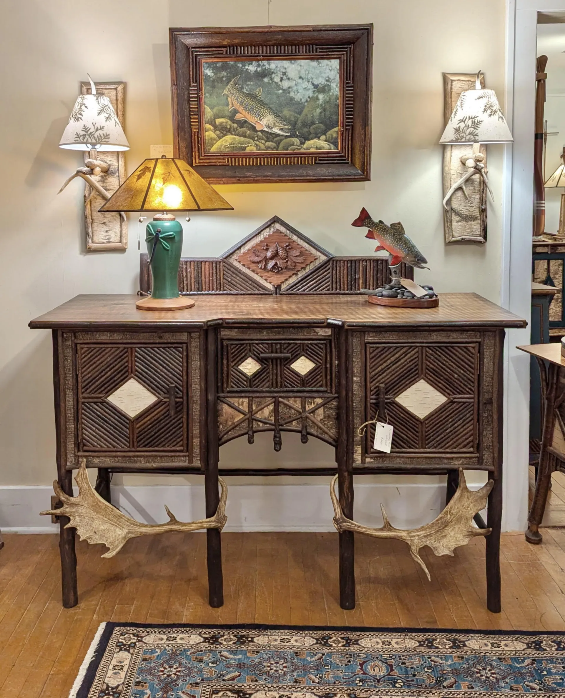 A rustic buffet table decorated with twig work and antlers, and a painting of salmon among a rocky stream bed in a matching twig frame.