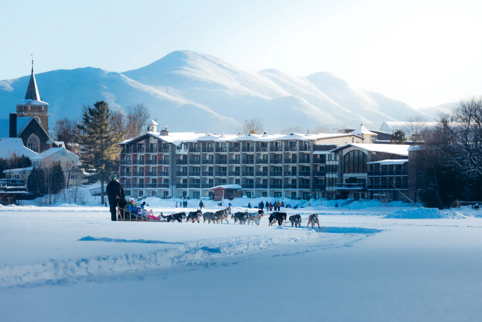 Dog sled team pulls sled across frozen lake with downtown on shoreline and snow-covered mountains in the background