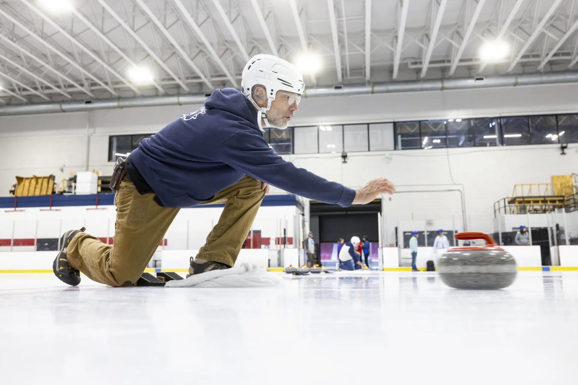 Man wearing long pants, sweatshirt, and helmet kneels on one knee on the ice after pushing away curling stone