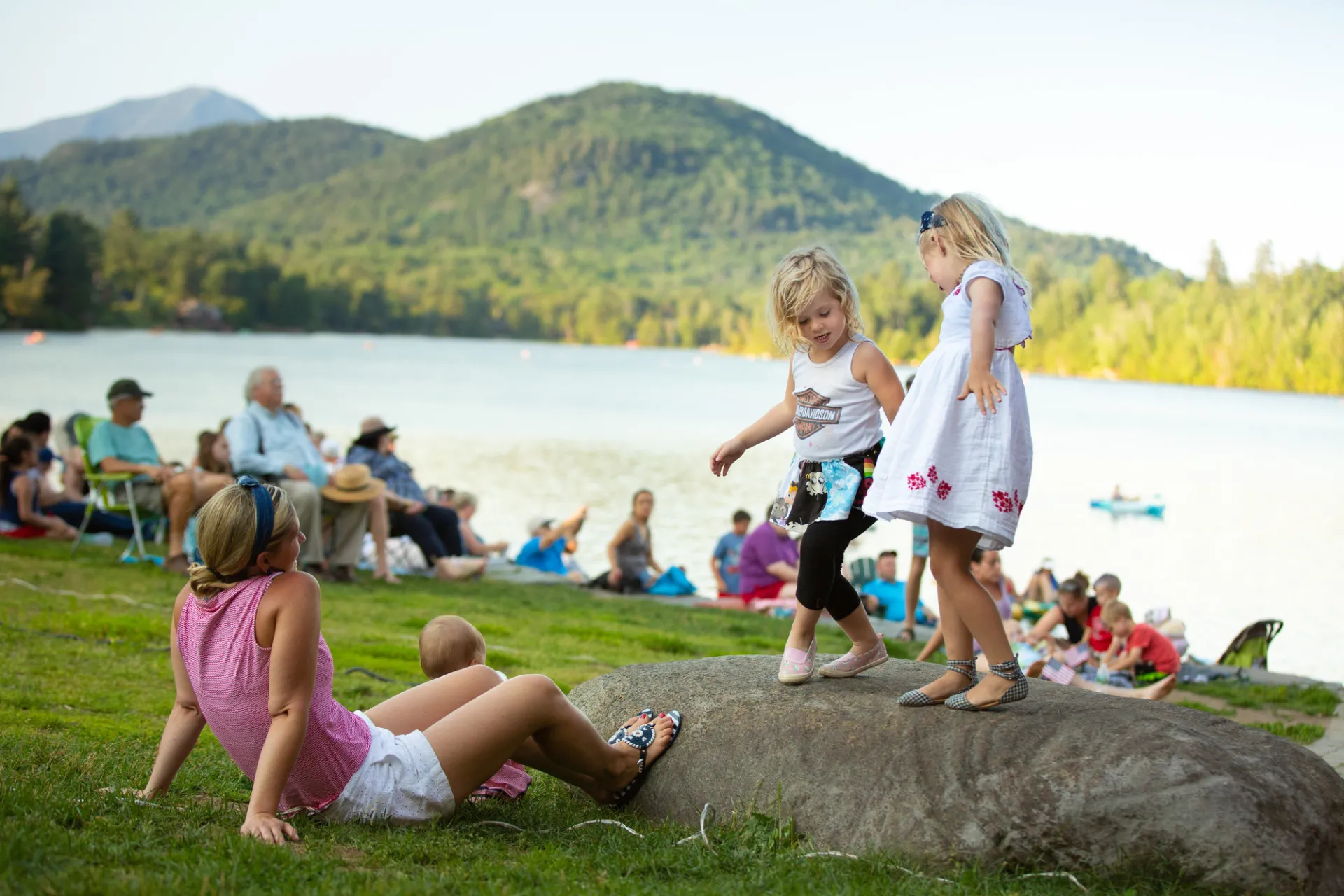 Two children play on a rock in a green park, with other people lounging on the lawn.