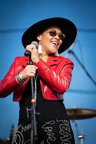 Dawn Tyler Watson on stage holding microphone waring red leather jacket, sunglasses, and black wide brim hat
