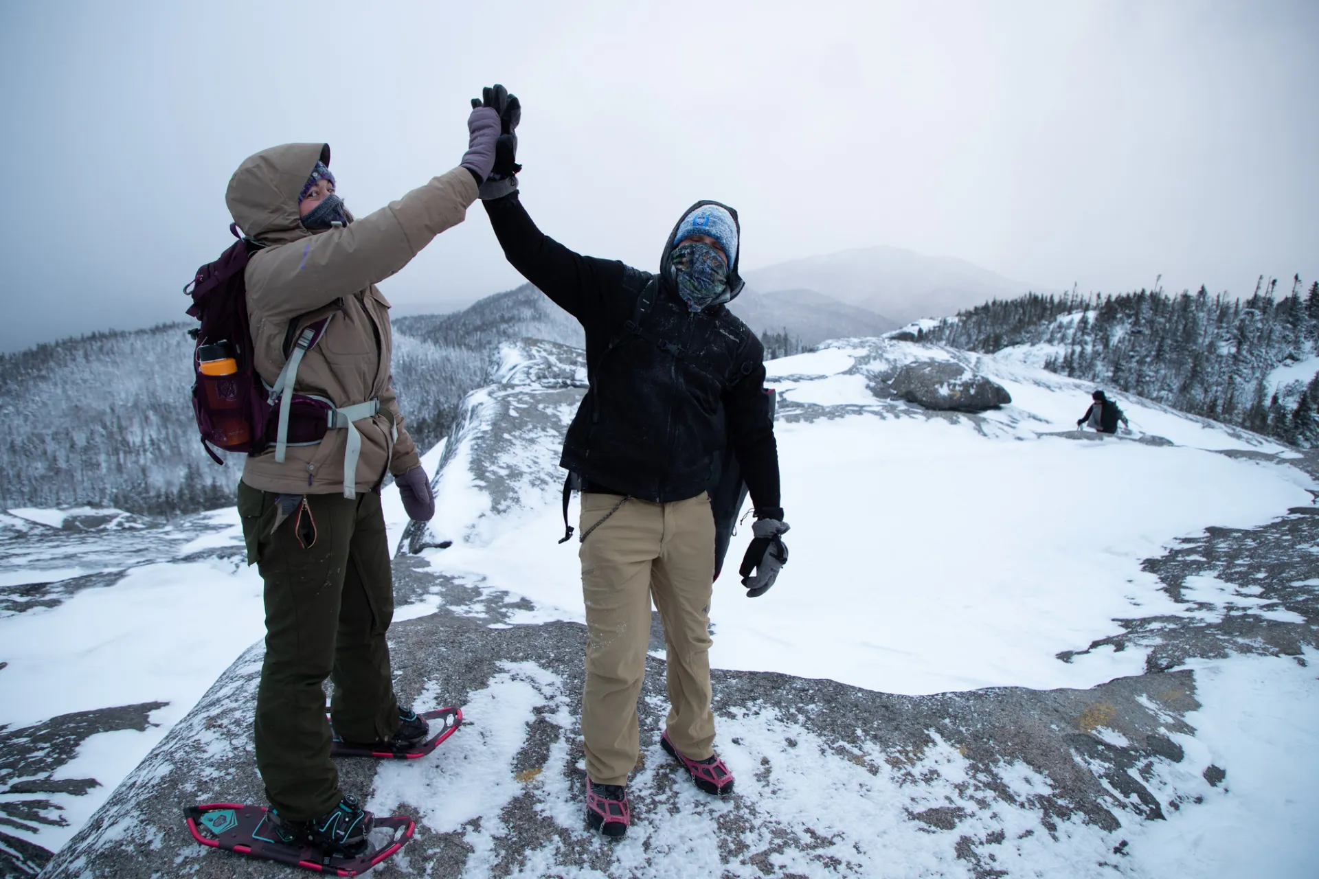 Two people wearing snowshoes and micro spikes high five at the summit of a wintry mountain.