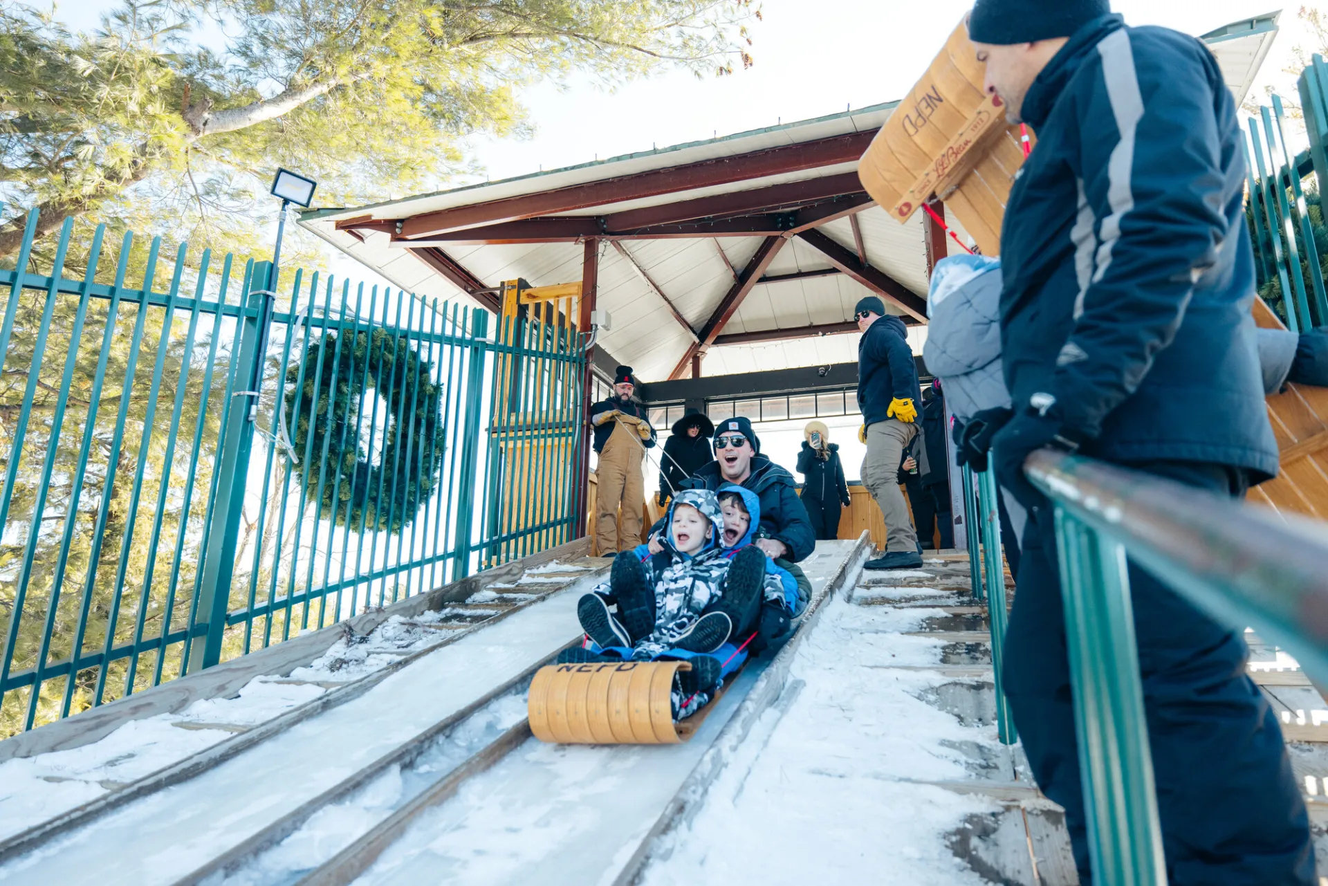 Man sits behind two children on a wooden toboggan, all wearing winter outdoor gear. 