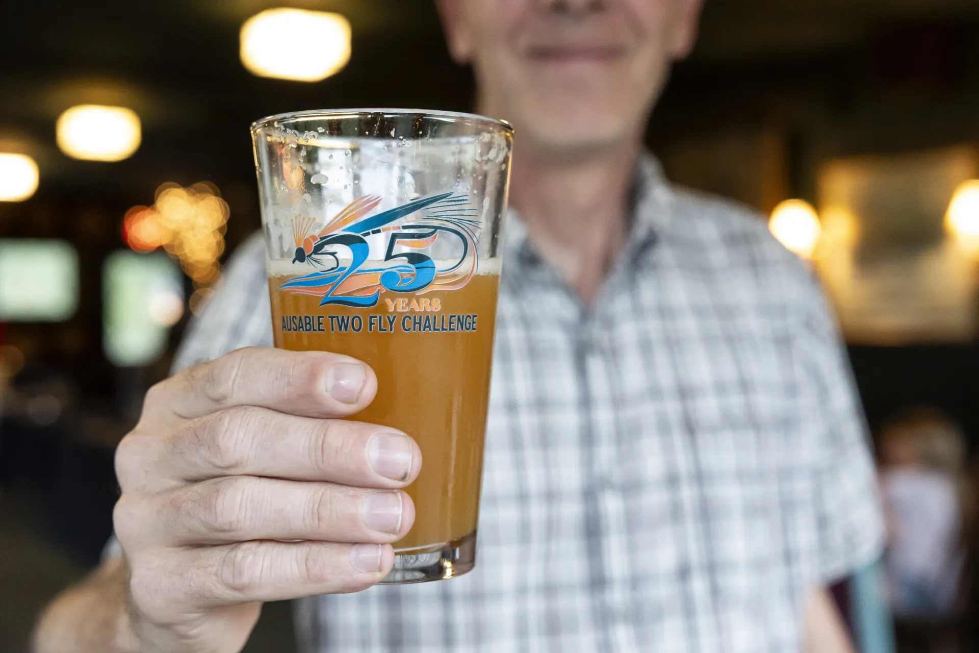 Man stands holding out 25th anniversary Ausable River Two-Fly Challenge pint glass filled halfway with beer