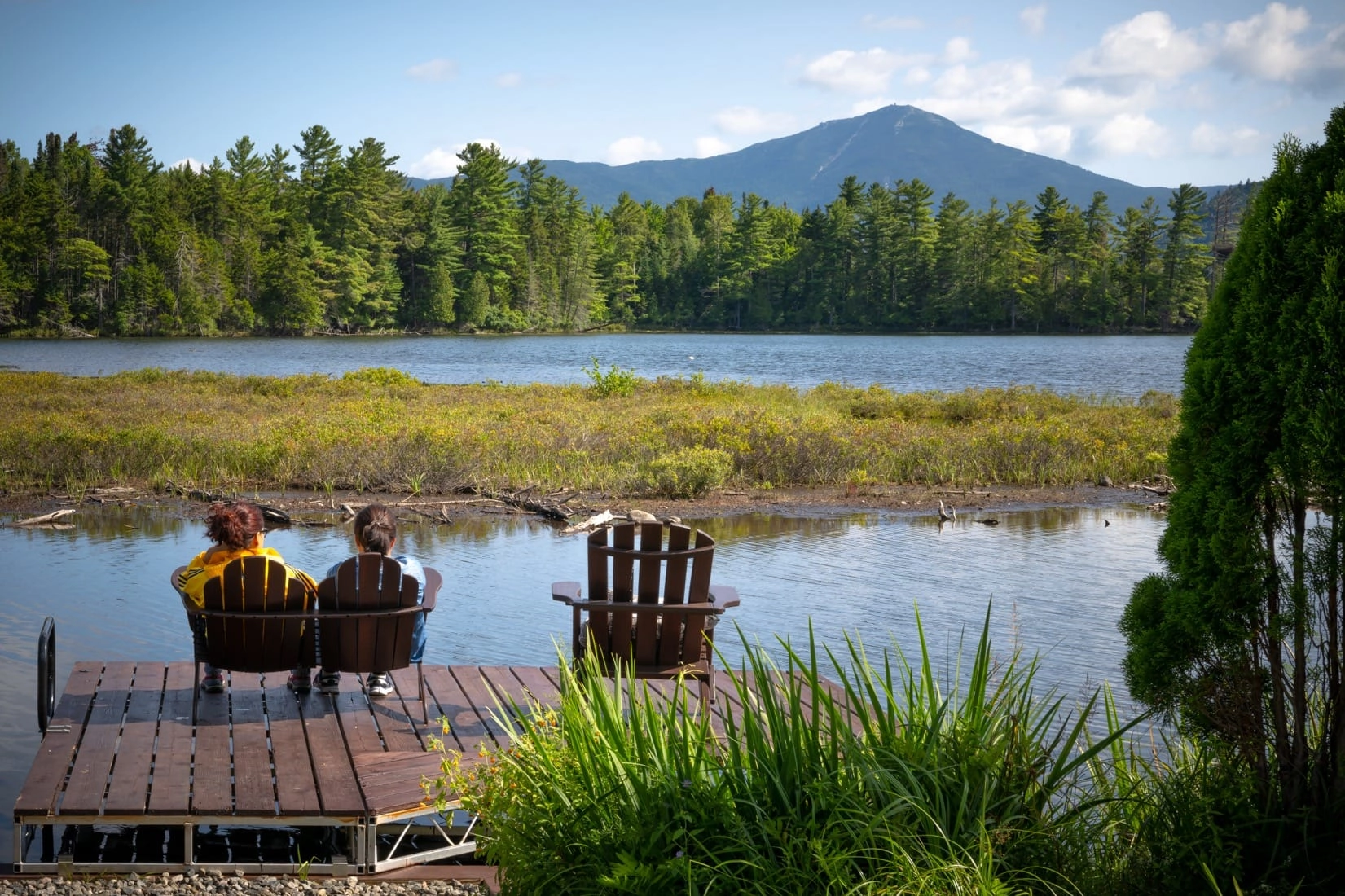 People sit in chairs on the dock by the water looking at the mountains.