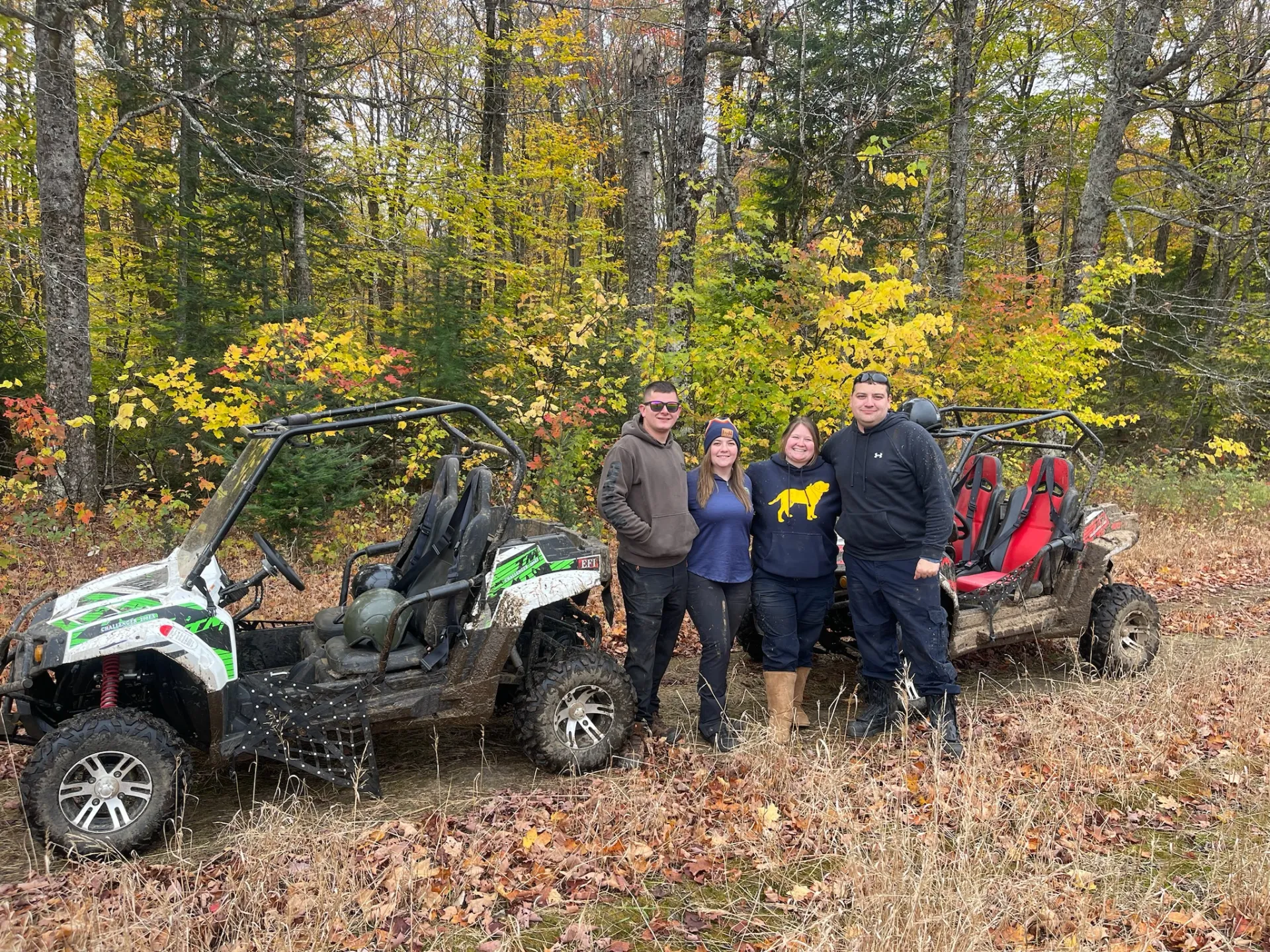 Four people stand together between two muddy UTVs surrounded by fallen leaves and vibrant fall foliage