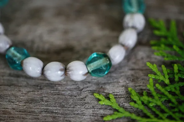 A close-up of a beaded bracelet with white and teal oval beads on a wooden surface, next to green fern-like leaves.
