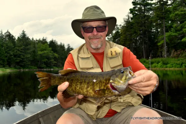 A man in a boat with a wide brimmed hat, sunglasses, a multi-pocketed vest and displaying a caught fish.