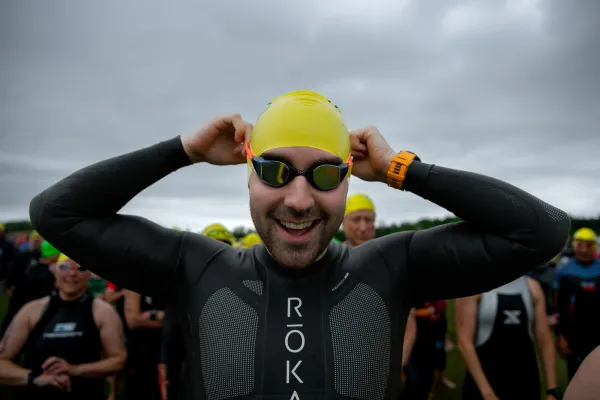 A smiling triathlete in a black wetsuit adjusts his goggles and yellow swim cap while preparing for the start of the race.