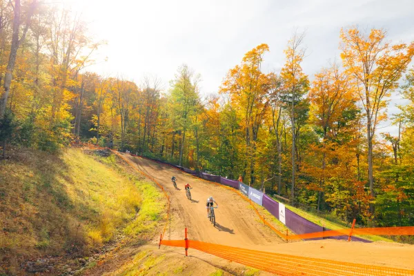 Mountain bikers ride down dirt trail on race course past fall foliage trees and event fencing