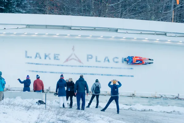 Luge racer rides down track with Lake Placid in ice while crowd looks on