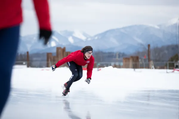 Skater in red coat crouches and skates on outdoor ice skating oval with mountains in the background and snow on the ground