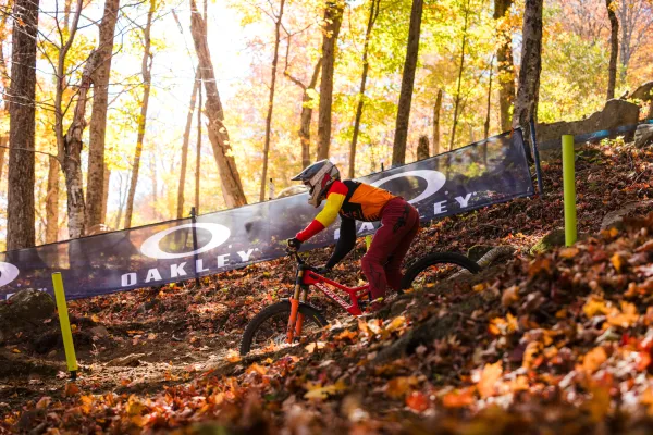 Downhill mountain bike races passes Oakley branded event fencing in forest with fall leaves during UCI MTB Dowhill World Cup