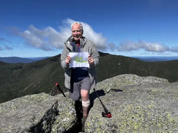 Older Gentleman stands atop a mountain peak holding a sign that reads '46er!'.