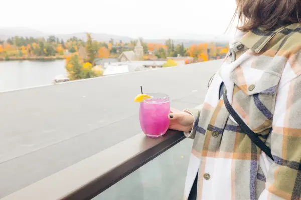 Woman in light flannel coat and painted fingernails holds glass of purple cocktail on rooftop bar overlooking Lake Placid in fall