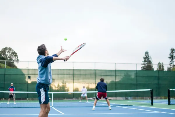 Man serves a tennis ball to an opponent on a tennis court beside another court where two players volley