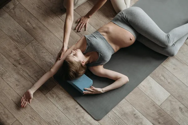 Pregnant woman in athletic clothing stretches on grey yoga mat on wooden floor with another person's arms helping deepen the stretch