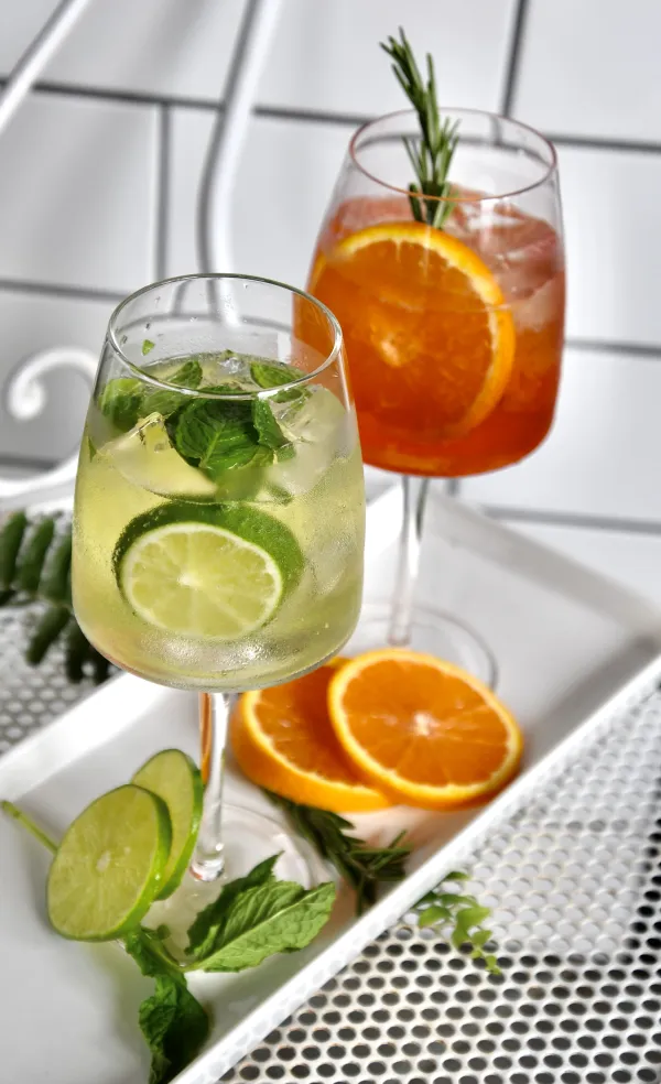 Two cocktails that look like a mojito and an Aperol spritz with a rosemary sprig on a white plate with citrus garnishes