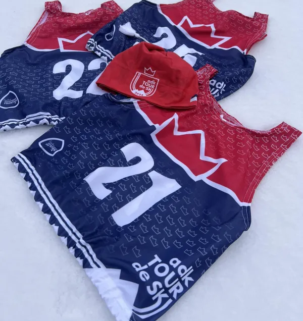Red and blue sports jerseys with a crown design lay on a snow surface. The number 21 is prominent, and a matching red hat with "Tour de Ski" logo is placed in the center.