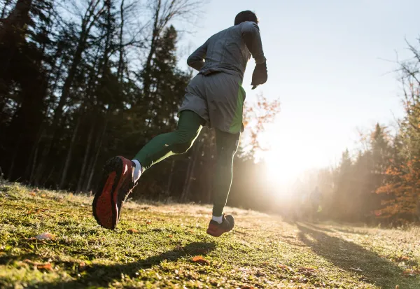 Close up of person's feet as they run on grassy path toward sunlight with bare and evergreen trees on sides of path