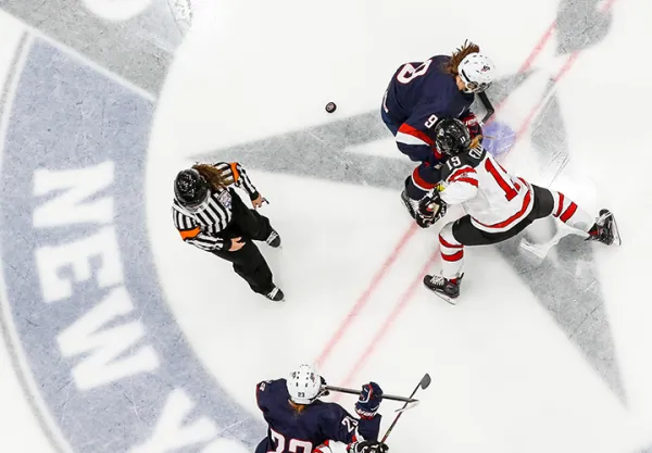 Two female hockey players face off on ice with another playing and a referee looking on