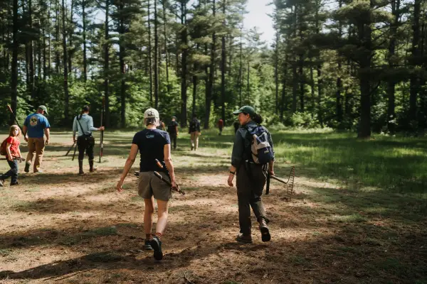 A group of volunteers walk away from the camera through a sunny forest clearing, carrying gardening tools.