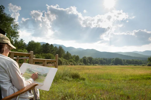 A person in a hat sketches a scenic landscape. The sun shines through clouds above mountains, a field, and trees. The mood is serene and creative.