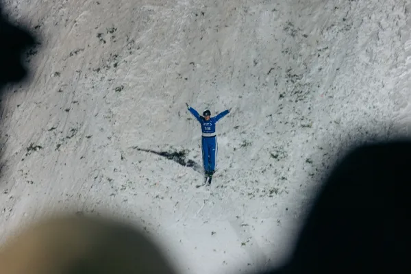 Freestyle aerial skier in a blue snowsuit lands on snowy hill sprinkled with pine bows. Their arms are extended out and up, while their legs  and skied feet are tight together