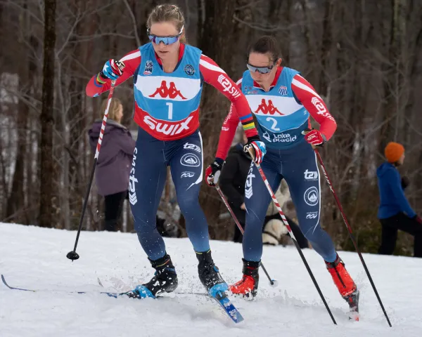 The image shows two female cross-country skiers racing on snow. 