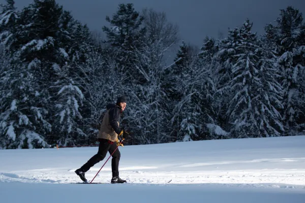Man in cross-country ski gear skis across open snow-covered filed with snow-covered pine trees behind him at night