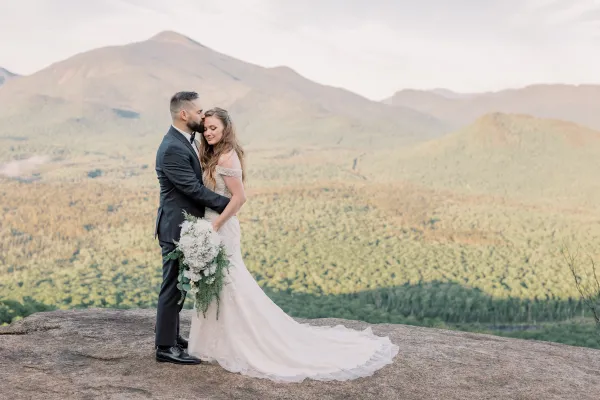 Groom kissses bride's forehead while she displays her bouquet on a rocky face with mountain views