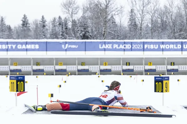 The image shows an athlete in a prone position, preparing to shoot during a biathlon event at the 2023 FISU World University Games in Lake Placid. 
