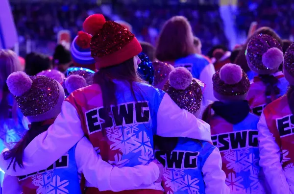 A group of people, mostly children, wearing festive red, white, and blue jackets with "ESWG" and snowflake designs, huddle together affectionately.