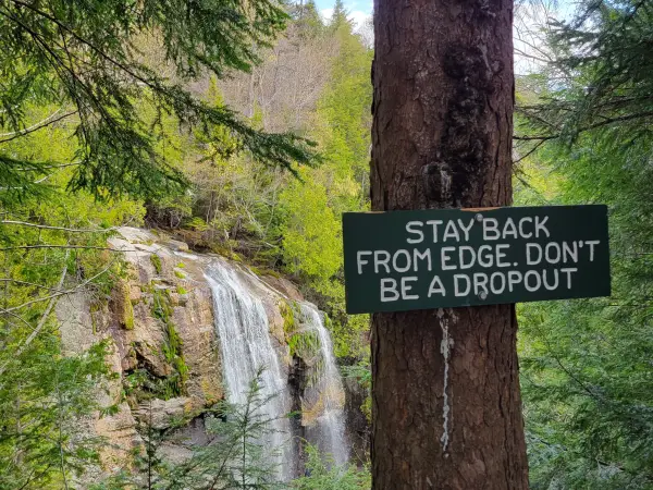 A sign that says "Stay back from edge, don't be a drop out" in front of a waterfall