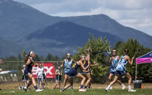 Two teams of young girls play lacrosse on green field with mountain backdrop.