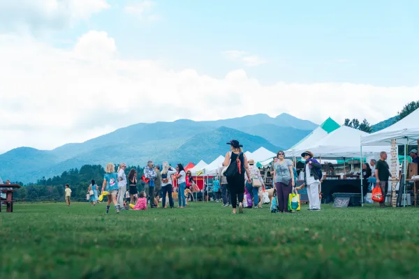 People browse white vendor tents at an outdoor market on a field, with mountains stretching across the horizon.