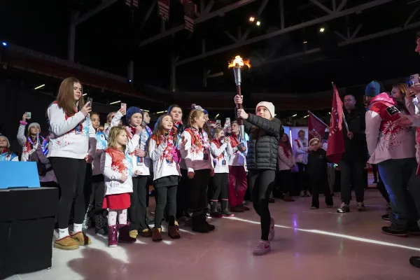 A young girl proudly carries a torch at the opening ceremony, surrounded by excited participants in ESWG jackets, capturing moments on their phones.