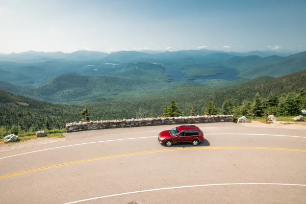 Red car drives on turn of high elevation highway road with mountain views and Lake Placid seen in the distance