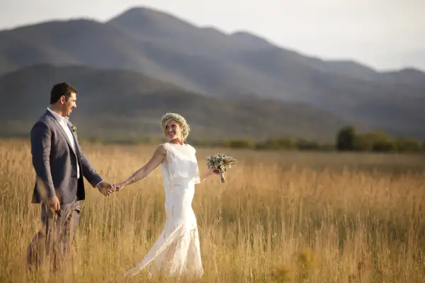 Bride and groom walk through a golden grass field with a mountain backdrop