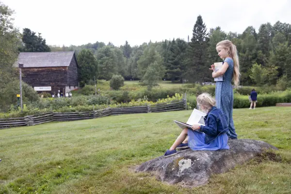 Girl sits on rock in dress and jacket reading book while second girl stands next to her holding book and looking out over green field with barn in distance