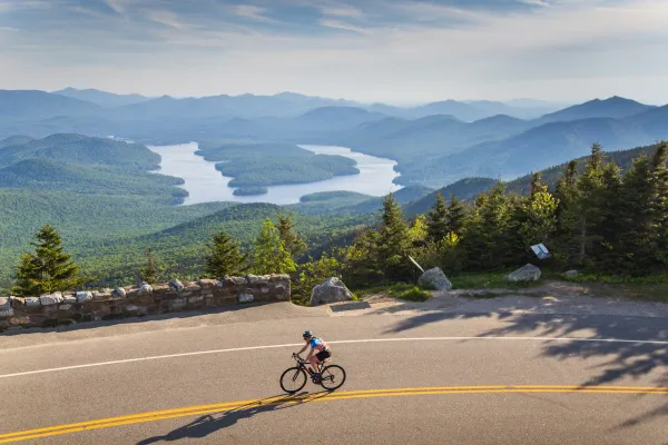 A cyclist rides along a mountain road overlooking a panorama of forested hills and an island-dotted lake in the valley below.