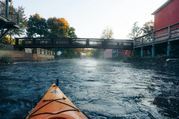 An orange kayak paddles down a misty river toward a low wooden bridge in the early morning light.