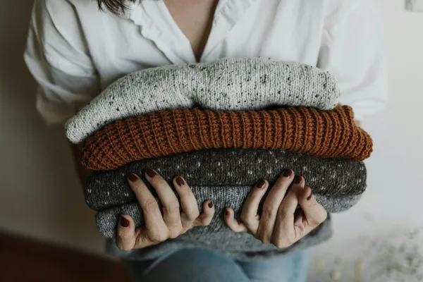 Person wearing white dress shirt and with nails painted a deep orange holds a stack of knit sweaters in her arms