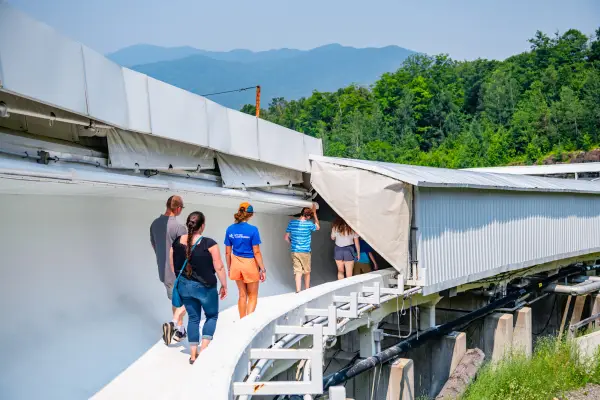 Family of five and female tour guide walk on bobsled track in summer with mountain views in background