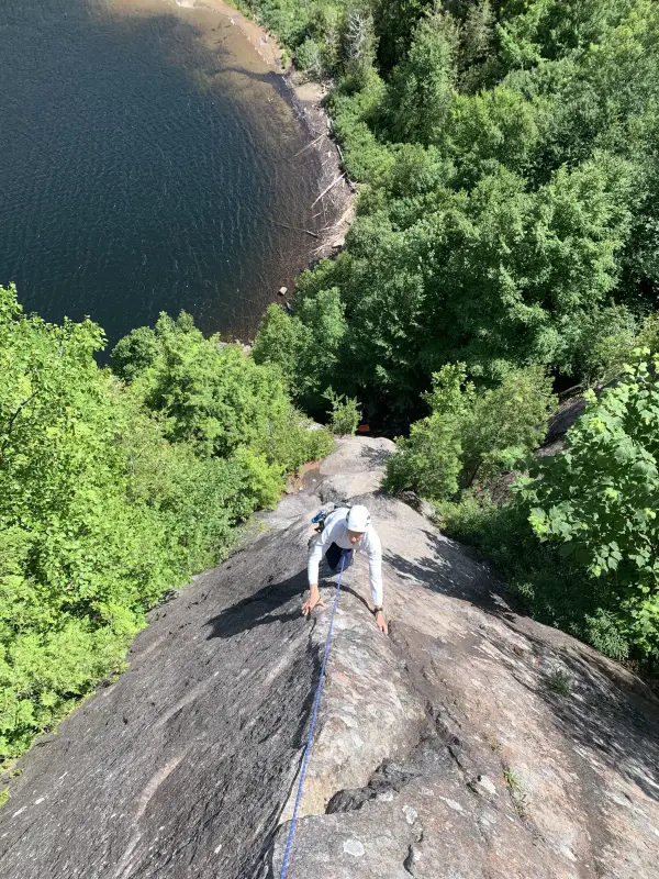 A rock climber ascends a steep cliff face overlooking a serene lake nestled within a lush green forest.