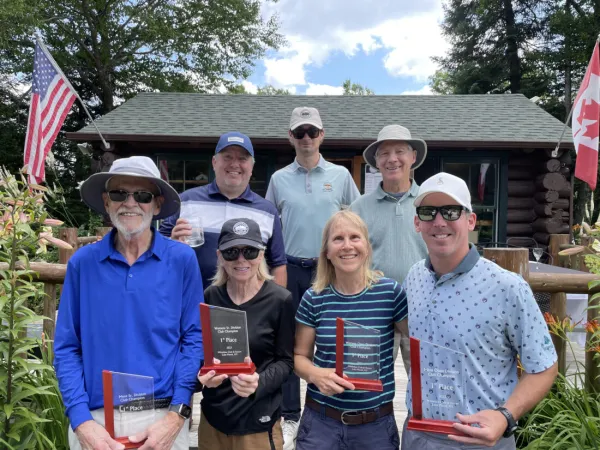 Seven people stand in two rows posing with glass awards from golf tournament outside on a summer day